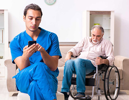 A male nurse sits in a chair using his mobile phone whilst an elderly patient calls out for help.